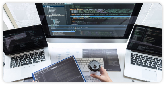 top view developer hand grabbing a cup in front of 2 laptop and 1 monitor 